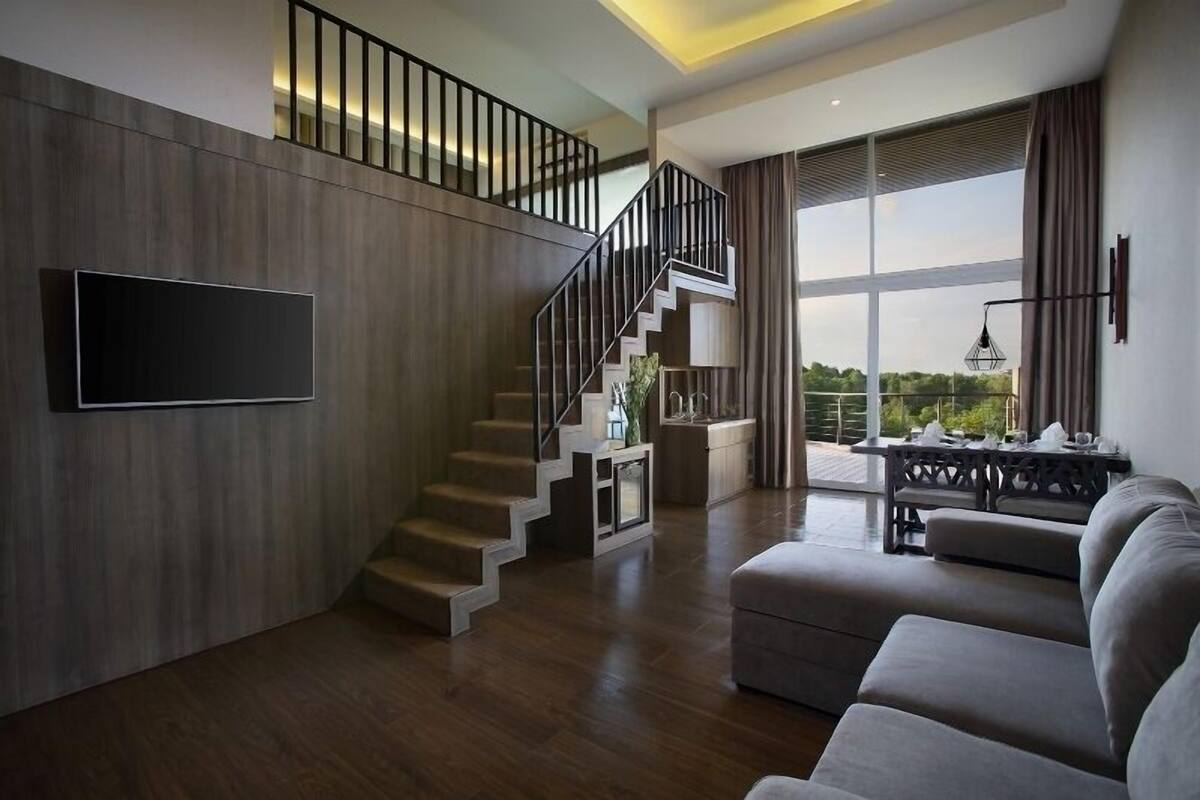 A modern living room with a staircase, a flat-screen TV, and a large window with curtains.
