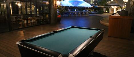 A pool table in a dimly lit indoor area with a bar and seating in the background.