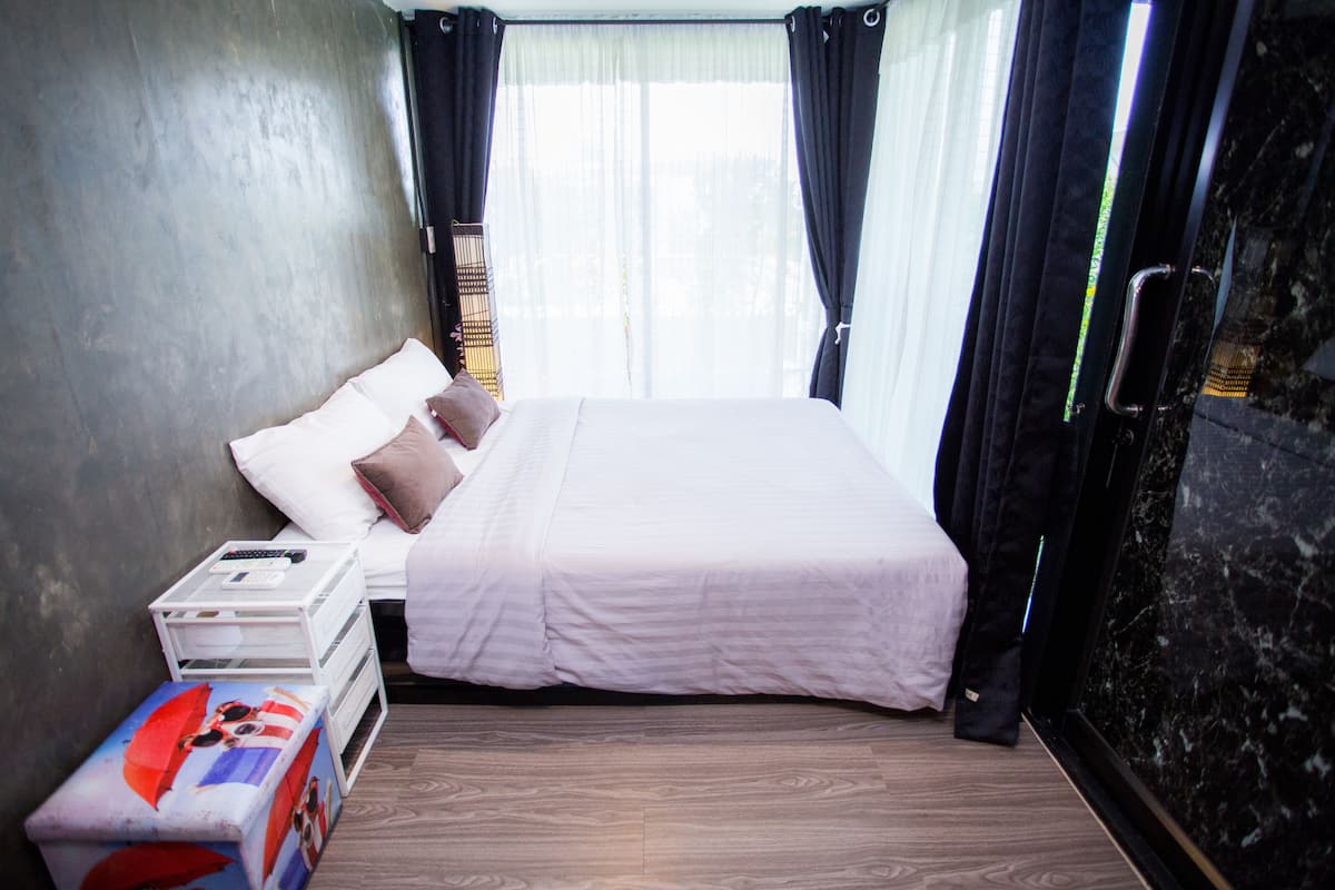 A bedroom with a bed, a bedside table with a remote control, a small table with a box, and a window with curtains.