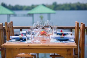 A wooden dining table set with glassware, plates, and a bowl of food, overlooking a lake with green umbrellas in the background.
