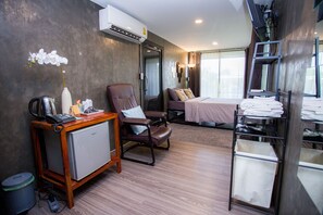 A hotel room with a bed, a desk with a computer, a chair, a small refrigerator, a coffee maker, a lamp, and a window with curtains.
