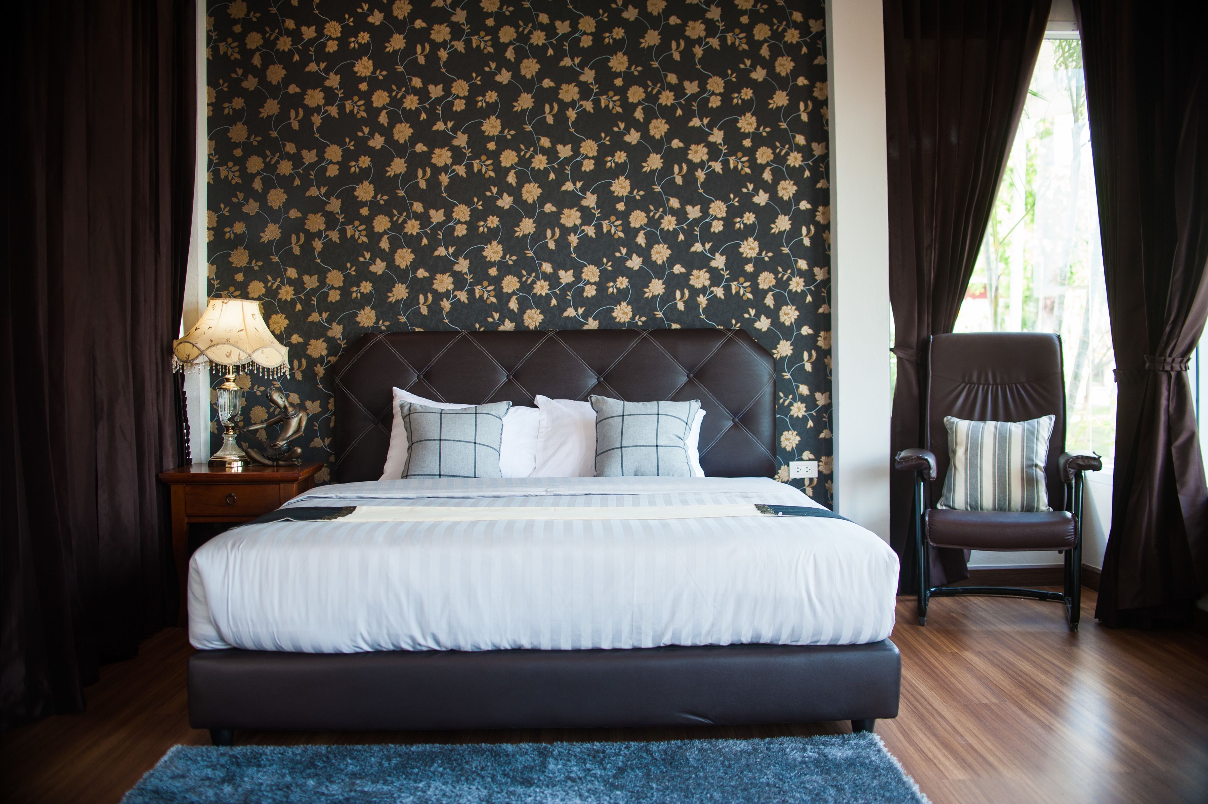 A bedroom with a large bed, dark curtains, a nightstand with a lamp, and a chair by the window.