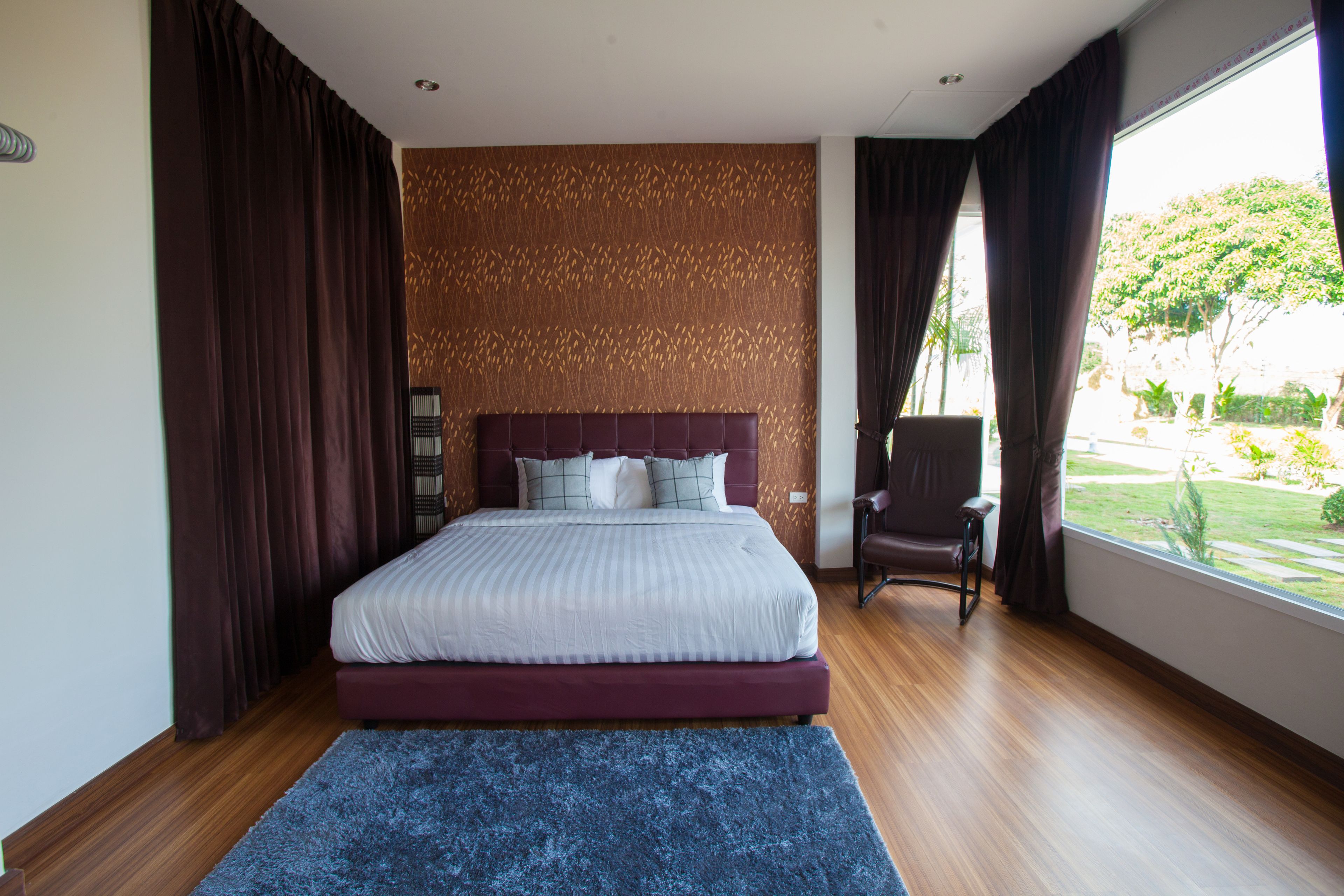 A modern bedroom with a large bed, dark curtains, a view of greenery outside, and a patterned wall.