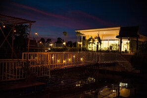 A nighttime view of a hotel with a well-lit entrance, a covered outdoor area with seating, and a bridge over a water feature.