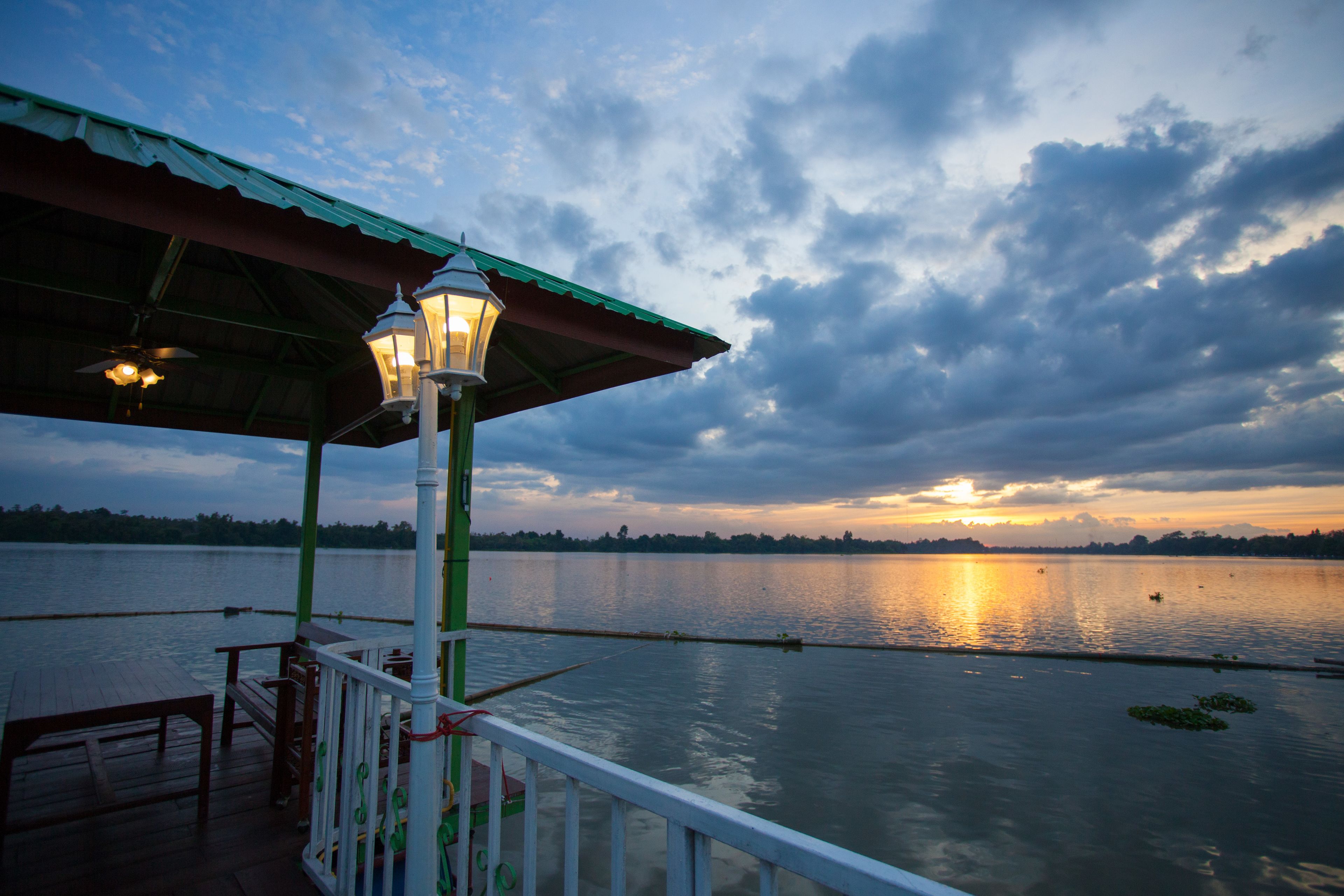 A wooden deck with a railing, a lamp post, and a covered area with a green roof overlooking a calm lake at sunset.