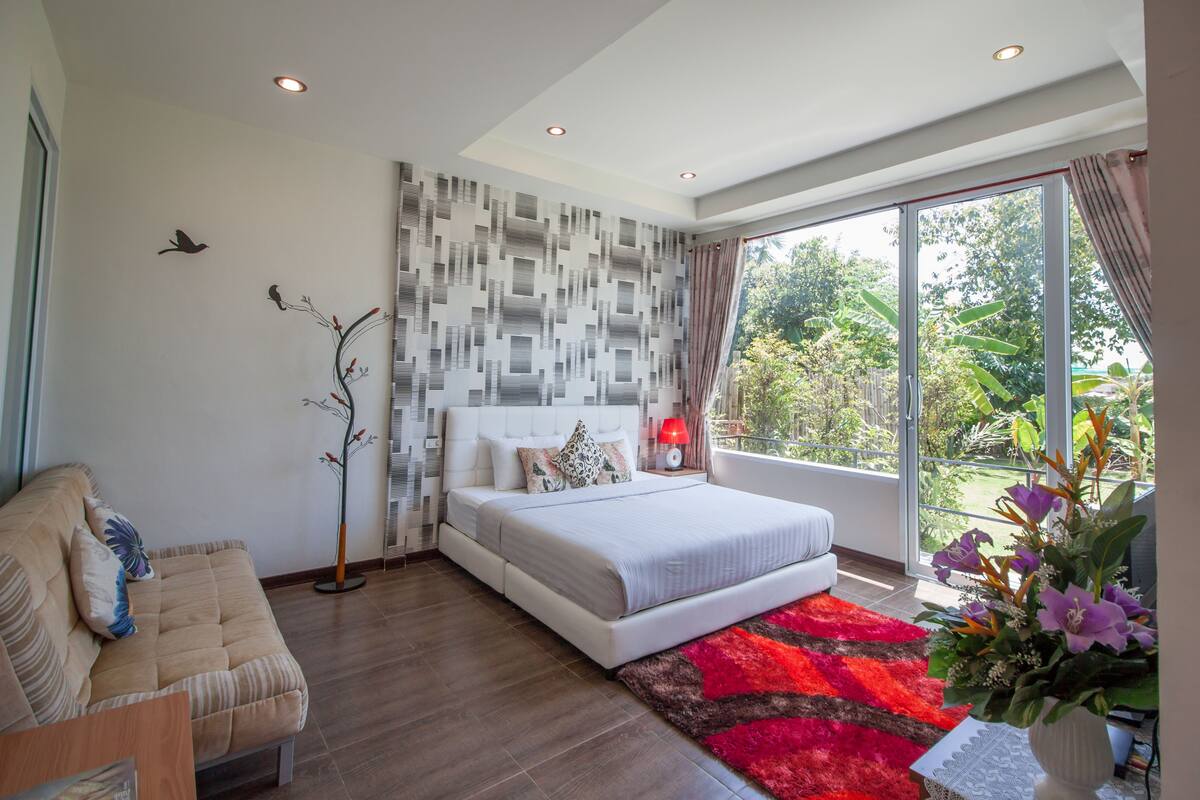 A modern bedroom with a large bed, a sofa, a floor lamp, and a view of greenery through the window.