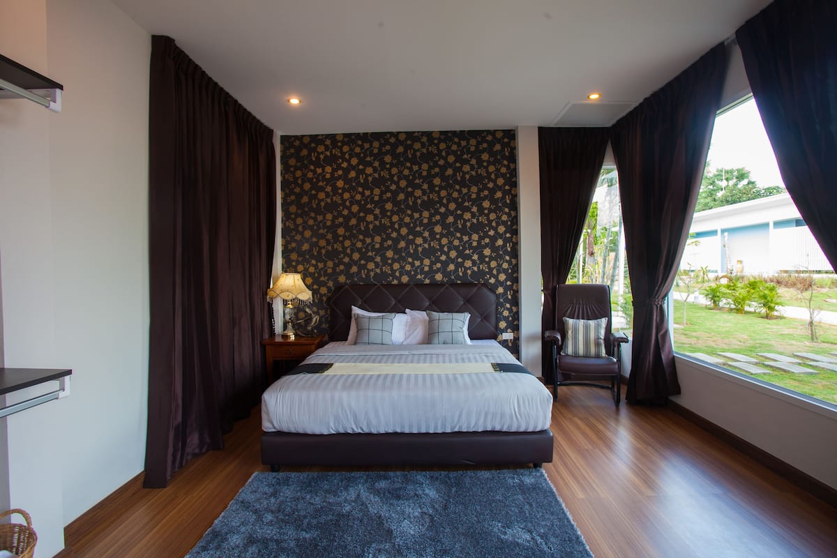 A bedroom with a large bed, dark curtains, a patterned wallpaper, a chair, and a view of the outdoors.