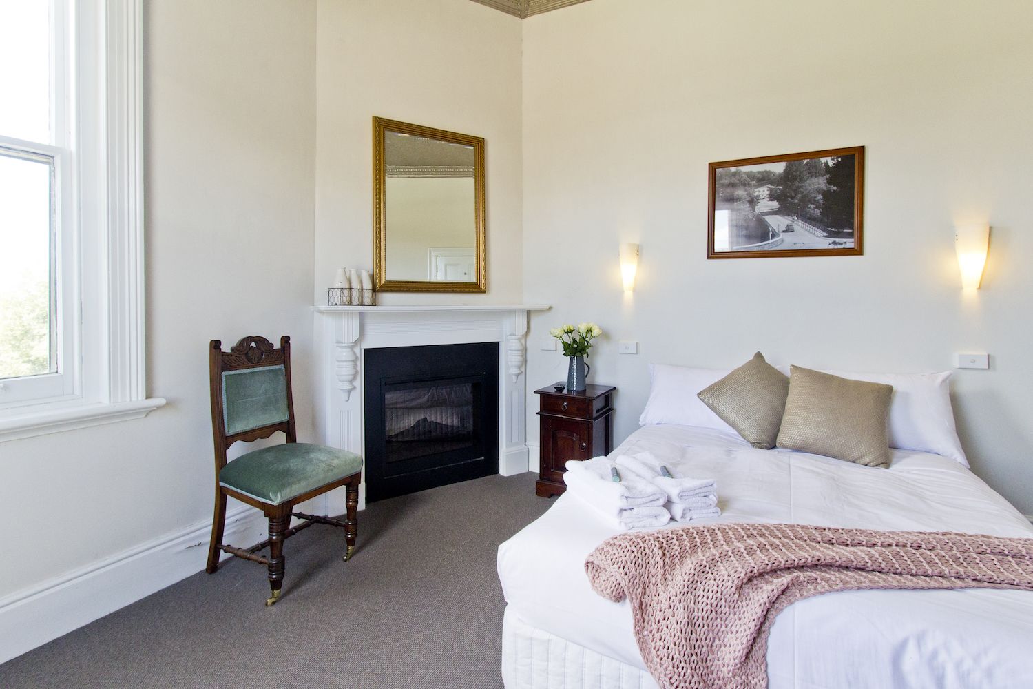 A bedroom with a fireplace, a bed with white linens, a green upholstered chair, and two wall-mounted sconces.