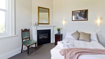A bedroom with a fireplace, a bed with white linens, a green upholstered chair, and two wall-mounted sconces.