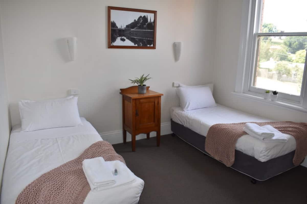 Ein Zimmer mit zwei Einzelbetten, einem kleinen Nachttisch mit Pflanze, einem gerahmten Bild an der Wand und einem Fenster mit Blick ins Grüne.