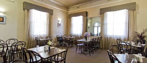 Comedor de hotel con varias mesas y sillas, ventanas amplias con cortinas y un techo con diseño texturizado.