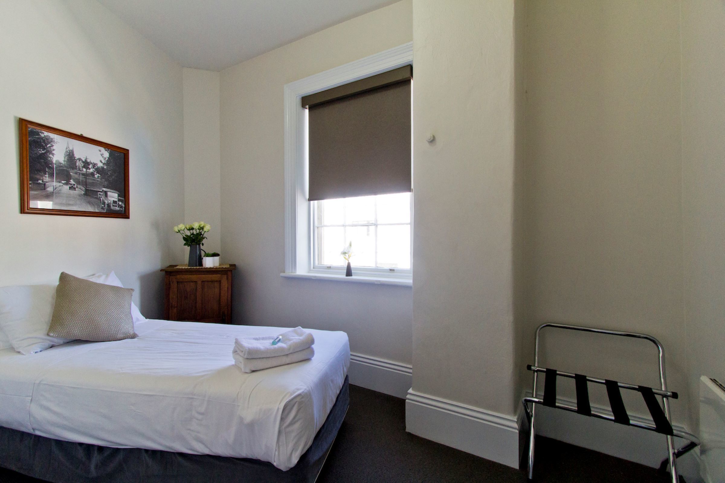 A bedroom with a bed, a window with a blind, a small wooden nightstand, and a framed picture on the wall.