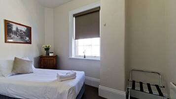 A bedroom with a bed, a window with a blind, a small wooden nightstand, and a framed picture on the wall.