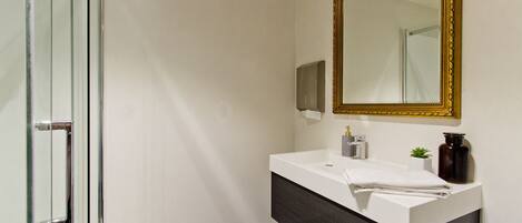 Un baño moderno con mampara de vidrio, un lavamanos blanco sobre un mueble de tocador oscuro y un espejo grande con marco dorado.