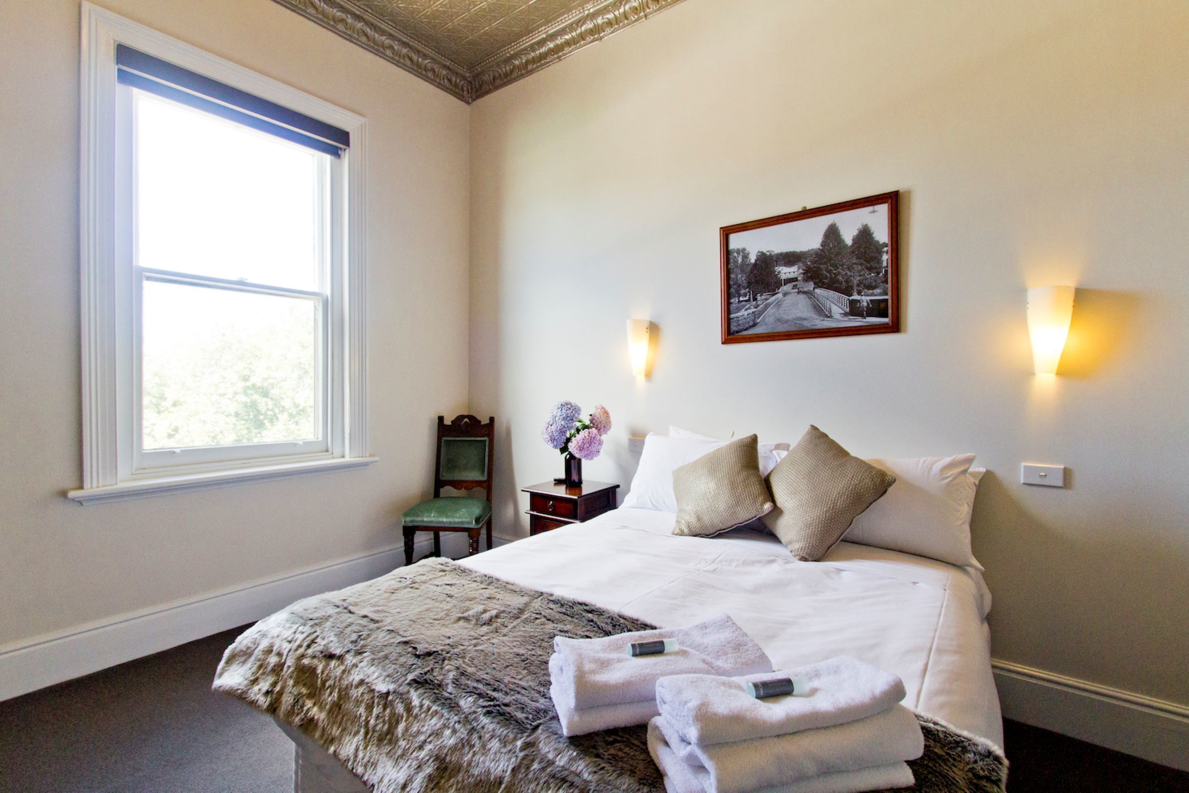 Un dormitorio con un ventanal grande, una cama con sábanas blancas, dos lámparas fijadas a la pared y un cuadro enmarcado.