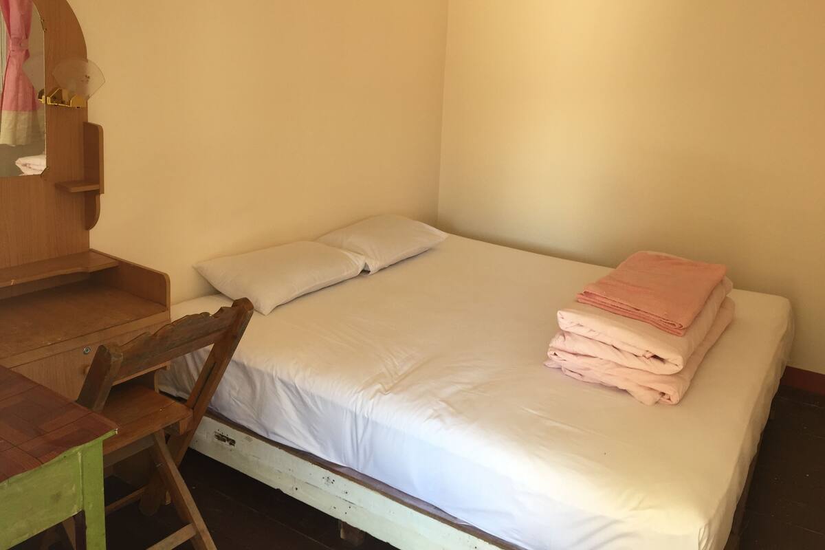 Un lit simple avec du linge de lit blanc et une serviette rose, un bureau en bois et une chaise dans une pièce aux murs unis.