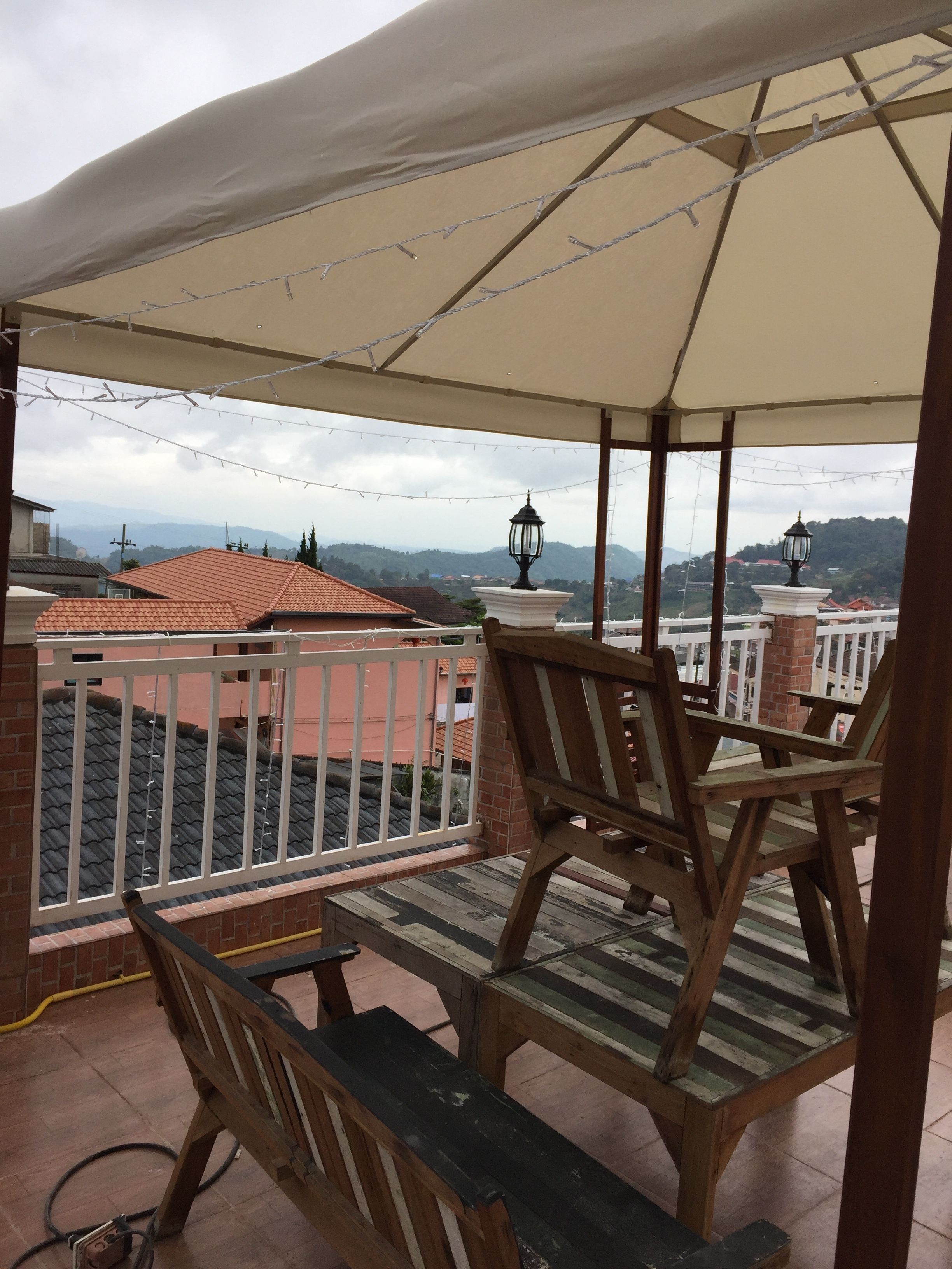 A covered outdoor seating area with wooden chairs and a table, overlooking a residential neighborhood with red-roofed houses and a hilly landscape in the distance.