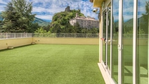 Un balcone con erba sintetica, vista su un castello su una collina e un edificio con ampie finestre.