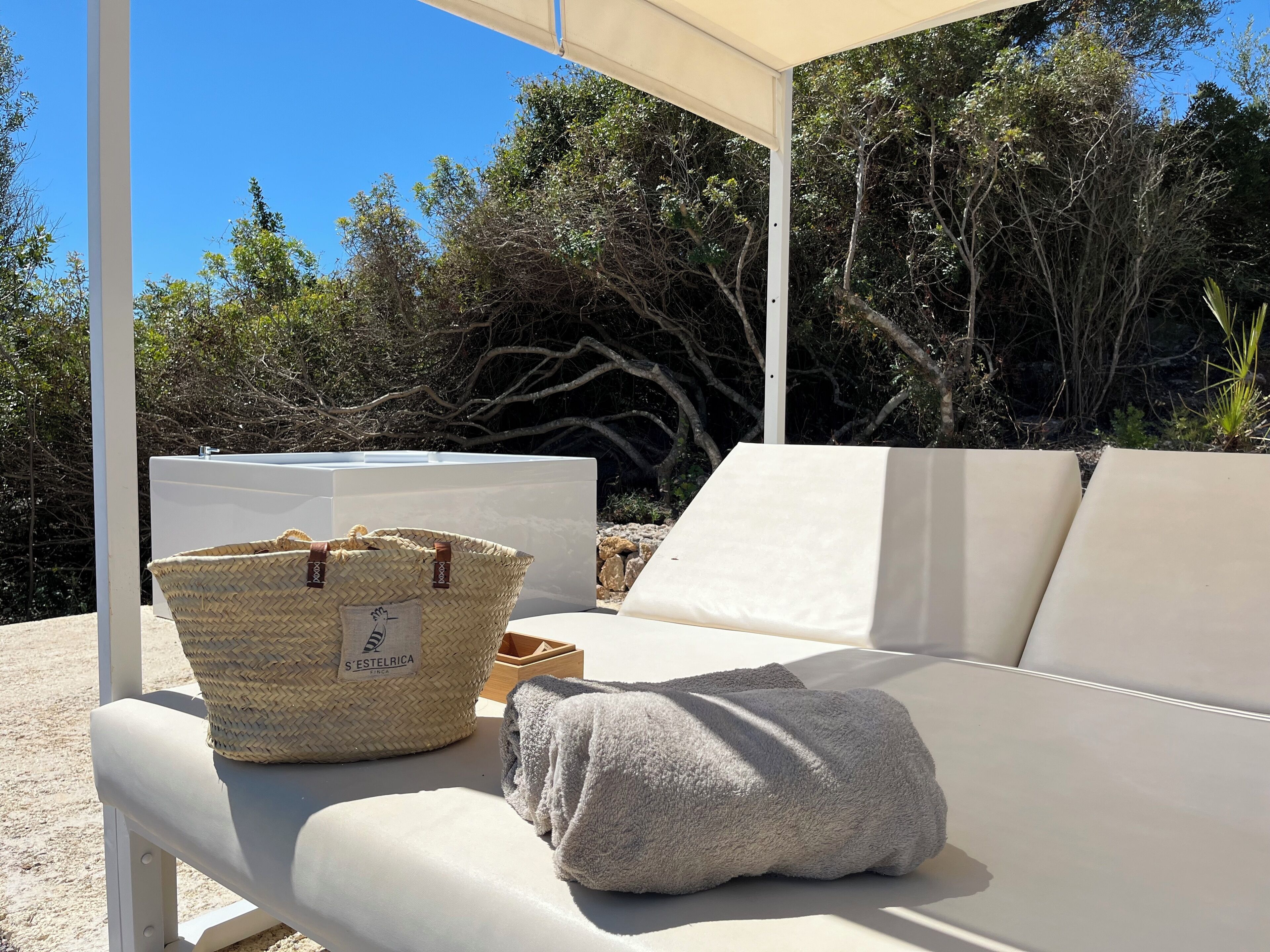 A sunlit outdoor seating area with a white cushioned bench, a woven basket, and a white container.