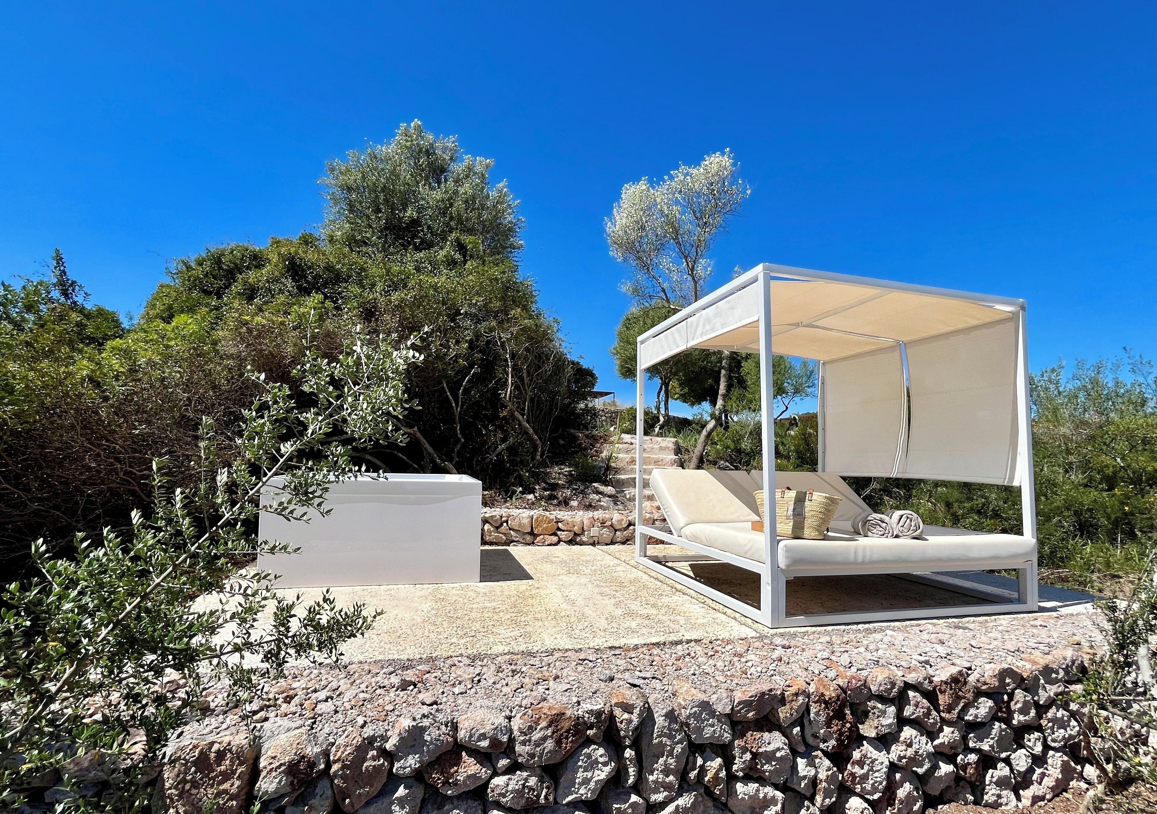 A white outdoor seating area with a canopy, a stone wall, and a view of trees and sky.