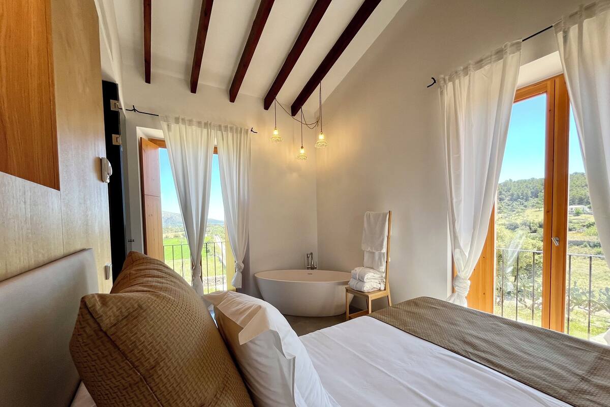 Quarto com uma cama grande, cabeceira de madeira, mesa de cabeceira e vista para a paisagem pela janela.