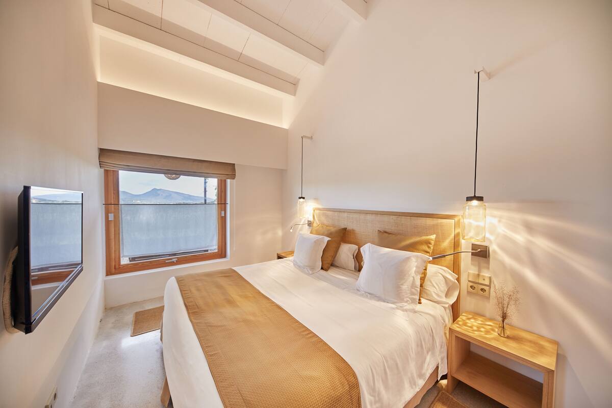 Quarto com cama grande, mesa de cabeceira, televisão montada na parede e janela com vista para montanhas.