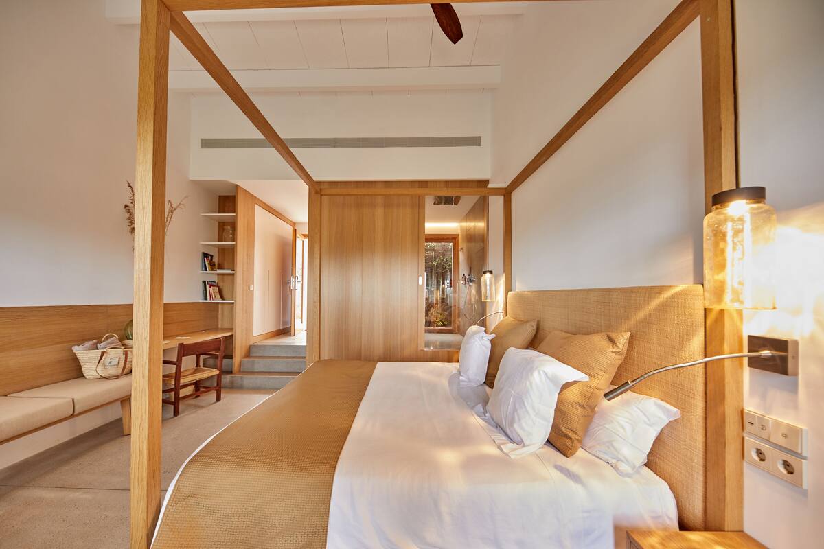 Quarto de hotel moderno com uma cama grande, cabeceira de madeira, mesa de cabeceira e uma prateleira embutida com livros e objetos decorativos.