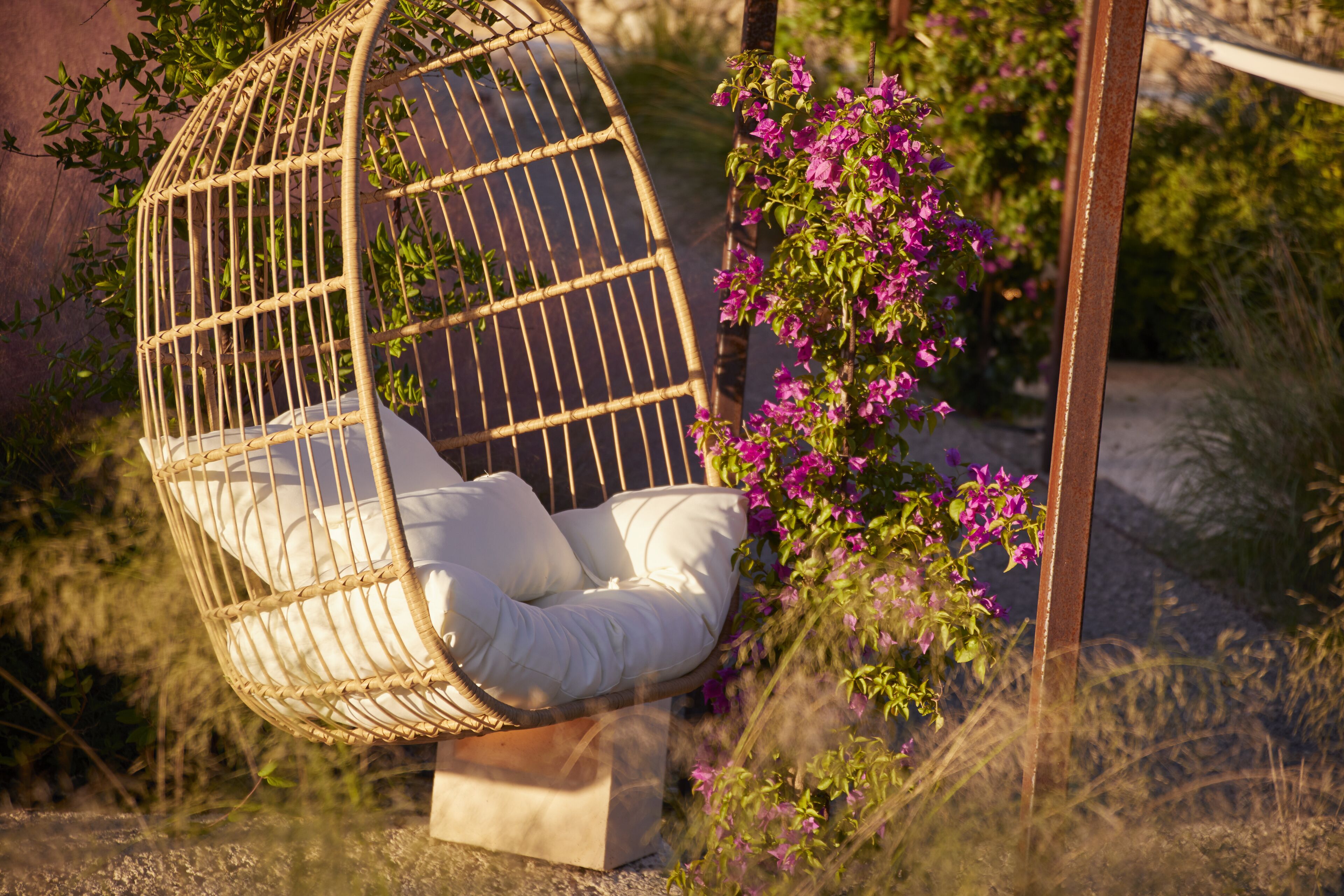 A wicker hanging chair with cushions, positioned outdoors with a view of greenery and flowers.