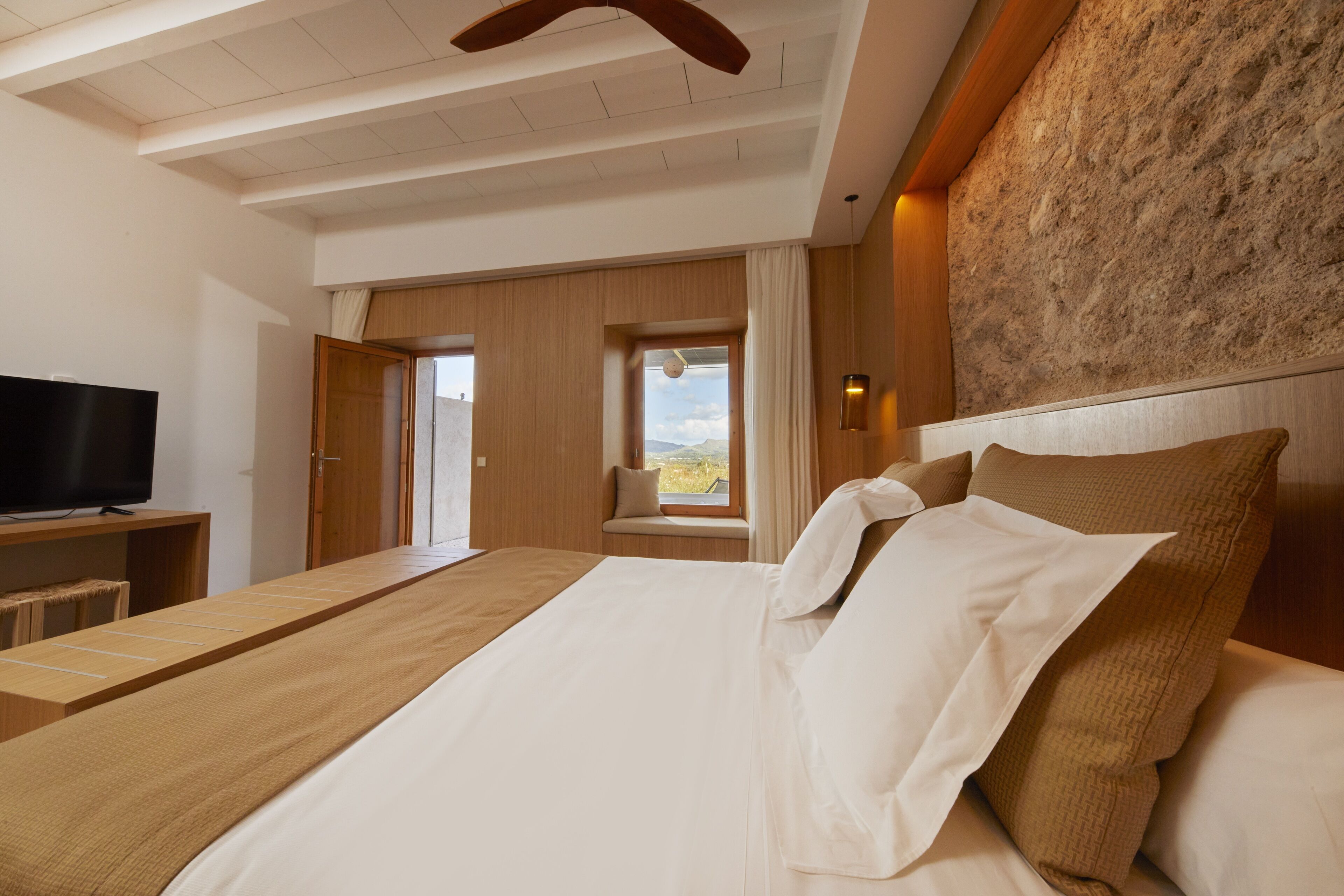A bedroom with a large bed, a television on a wooden stand, and a window with a view of the outdoors.