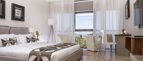 A modern hotel room with a large bed, a desk, and a view of the sea.