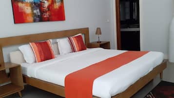 A neatly made bed with a wooden headboard, white and red pillows, and a red throw blanket. There is a small bedside table with a lamp and a vase with dried plants.