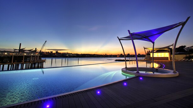 Una piscina infinita con terraza de madera, una estructura tipo toldo y un horizonte urbano al fondo.