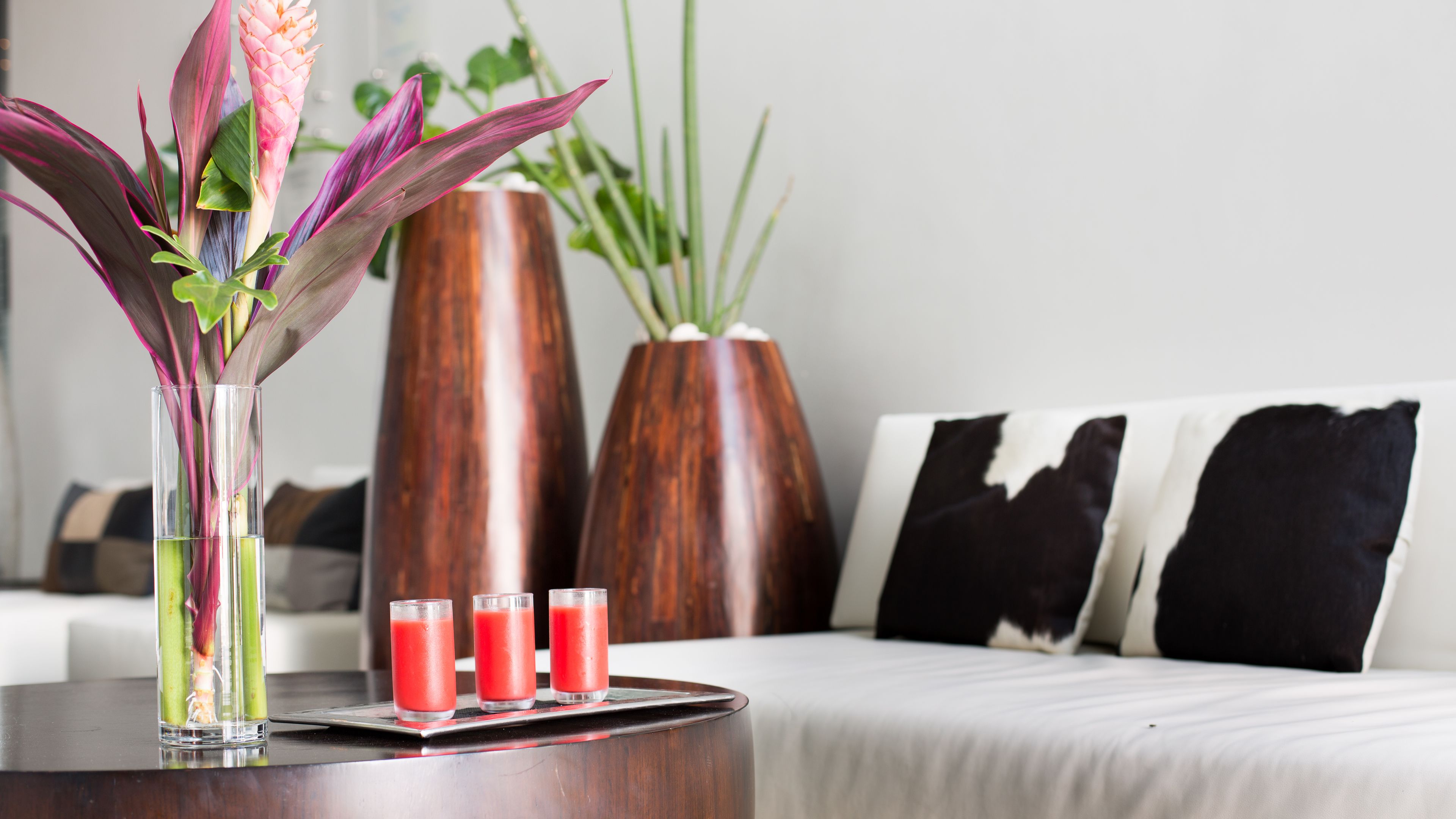 A modern living room with a coffee table holding a vase with flowers and three glasses of red beverage.