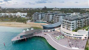 Vista aérea de un hotel frente al mar con un paseo marítimo, varios edificios de gran altura y una playa al fondo.