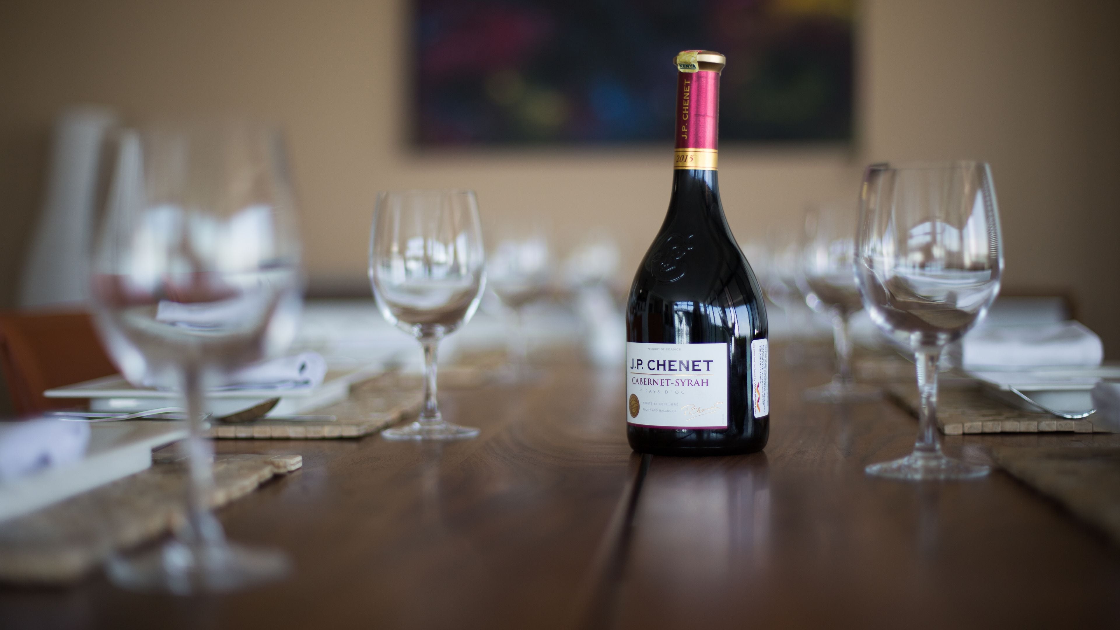 A bottle of J.P. Chenet Cabernet-Syrah on a wooden table surrounded by empty wine glasses.