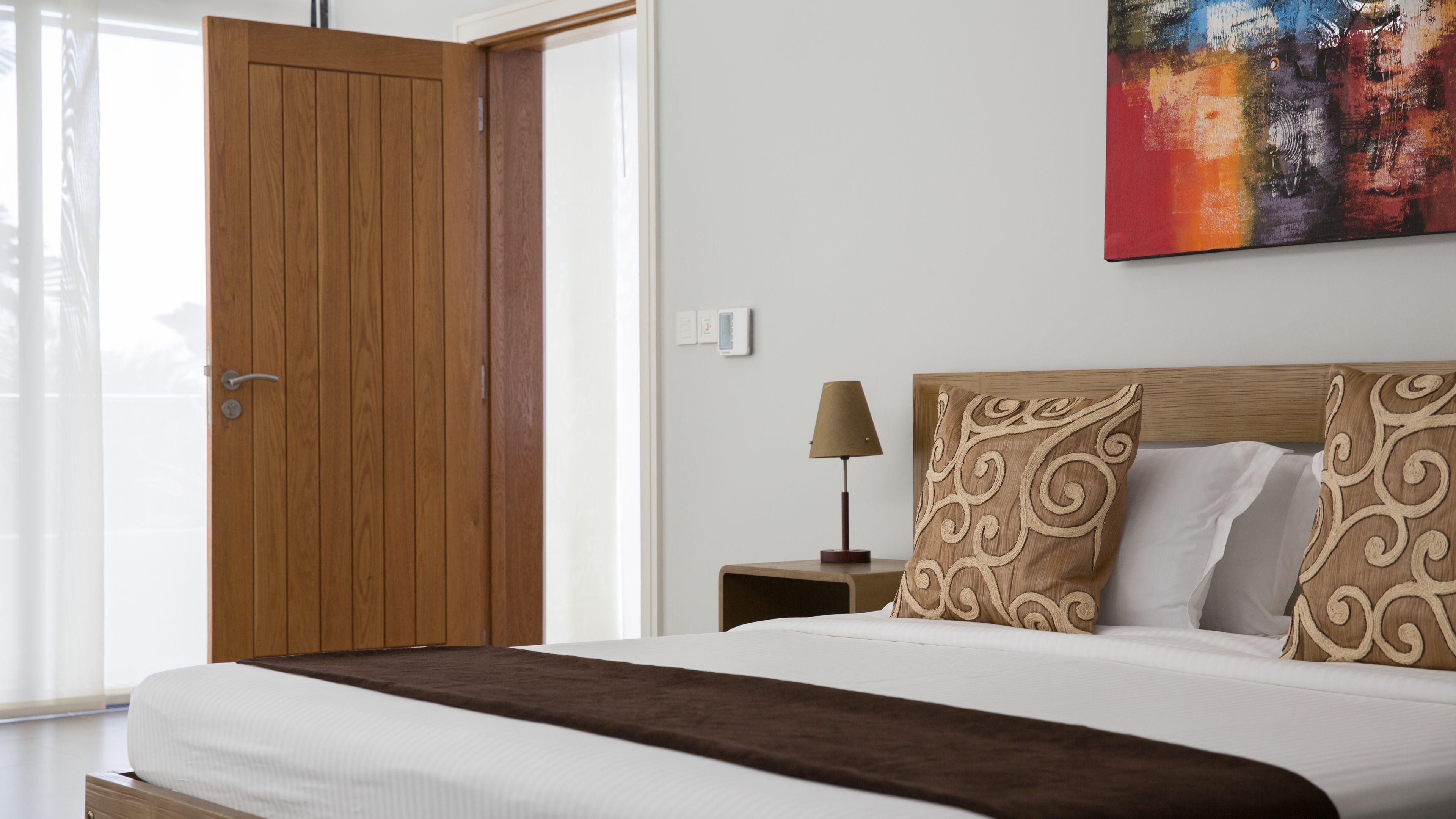 A bedroom with a wooden wardrobe, a bed with patterned pillows, a bedside table with a lamp, and a colorful abstract painting on the wall.