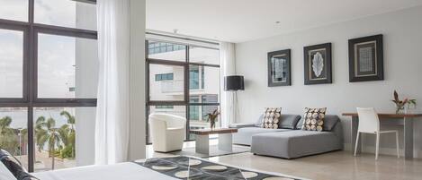 Una habitación de hotel moderna con una cama grande, un sofá gris, un sillón blanco y un comedor. Hay ventanas amplias con vistas a palmeras y edificios.
