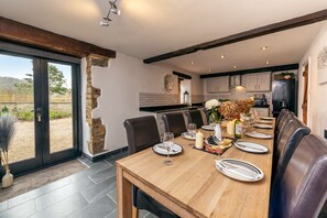 A dining area with a wooden table set for six, surrounded by leather chairs. A stone wall and a glass door lead to an outdoor area. - Somerset Country Escape (Taunton)