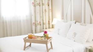 Una cama bien hecha con ropa blanca, una bandeja de madera con alimentos para el desayuno y una cortina floral al fondo.