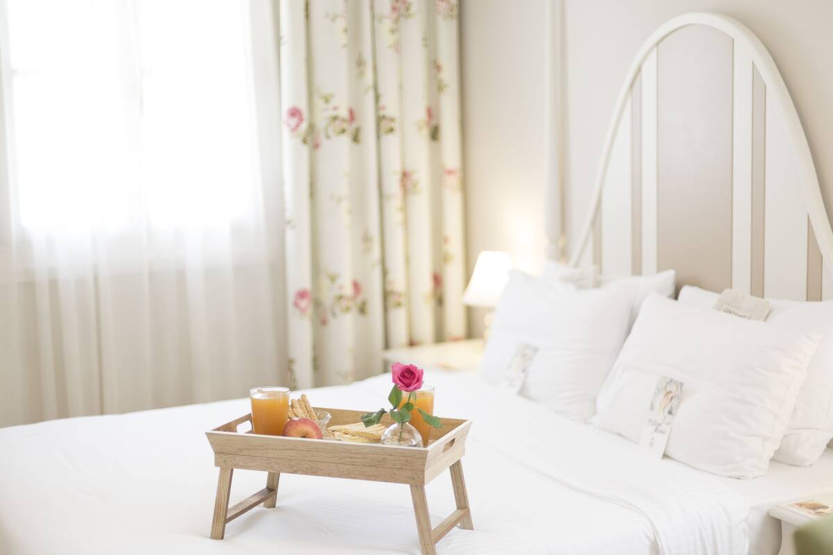A neatly made bed with white linens, a wooden tray with breakfast items, and a floral curtain in the background.