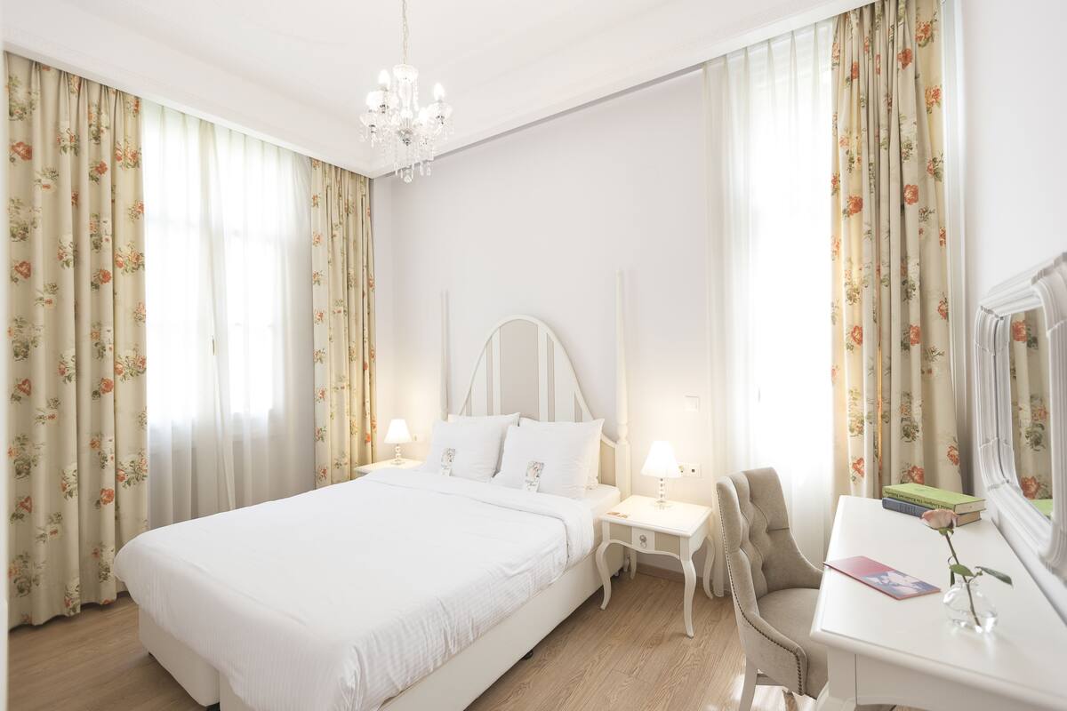 A hotel room with a large bed, a desk with a chair, a nightstand, and a chandelier.