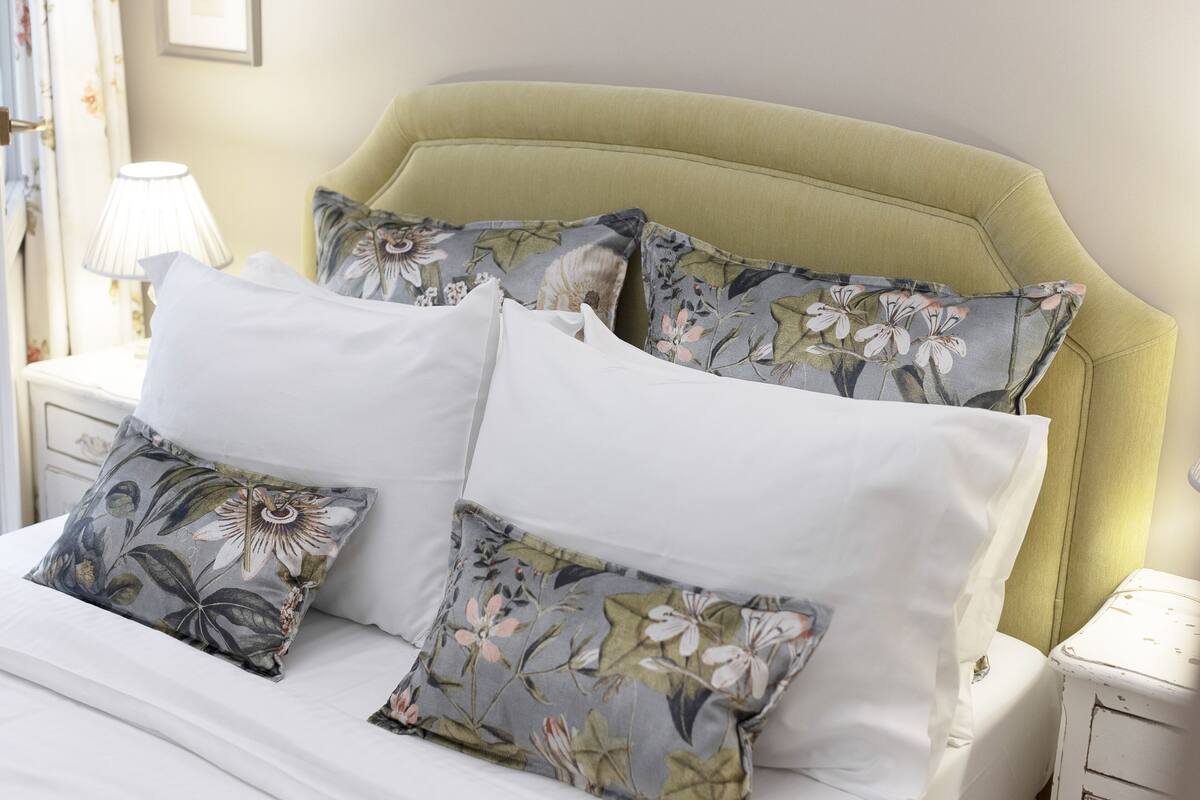 A bed with floral-patterned pillows and a plain white pillow, a bedside lamp, and a nightstand with a lamp.
