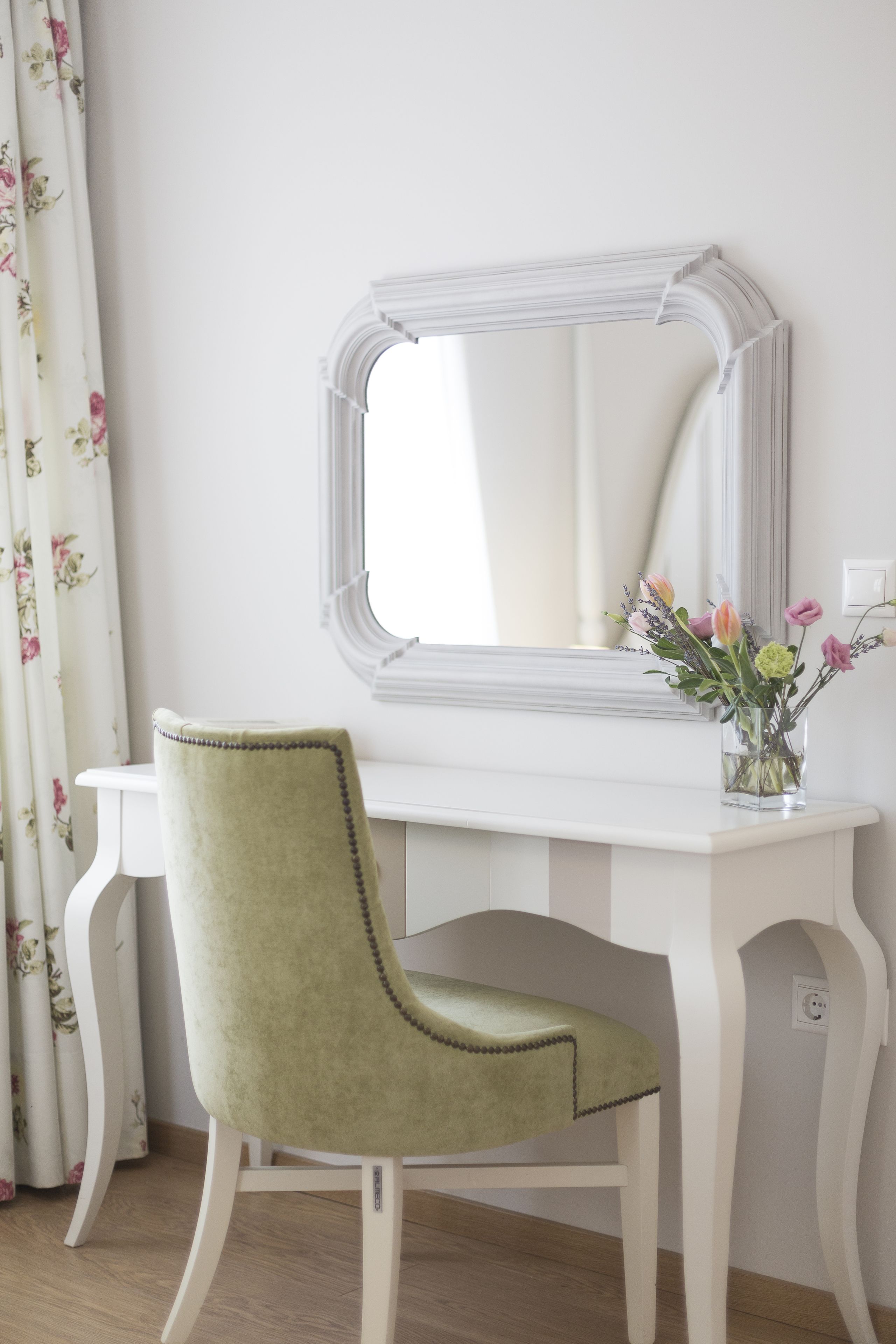 Une coiffeuse blanche avec un miroir, une chaise verte et un vase rempli de fleurs.