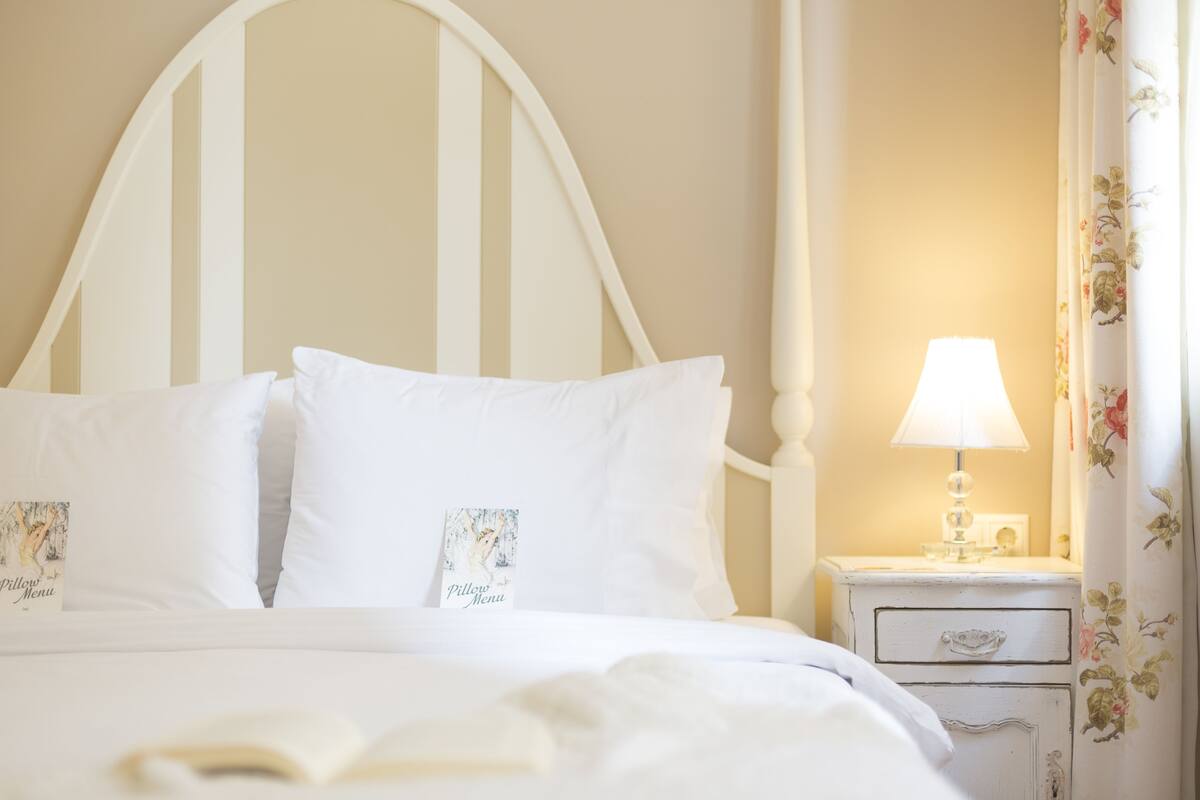 A bed with white bedding and pillows, a nightstand with a lamp, and a floral curtain.