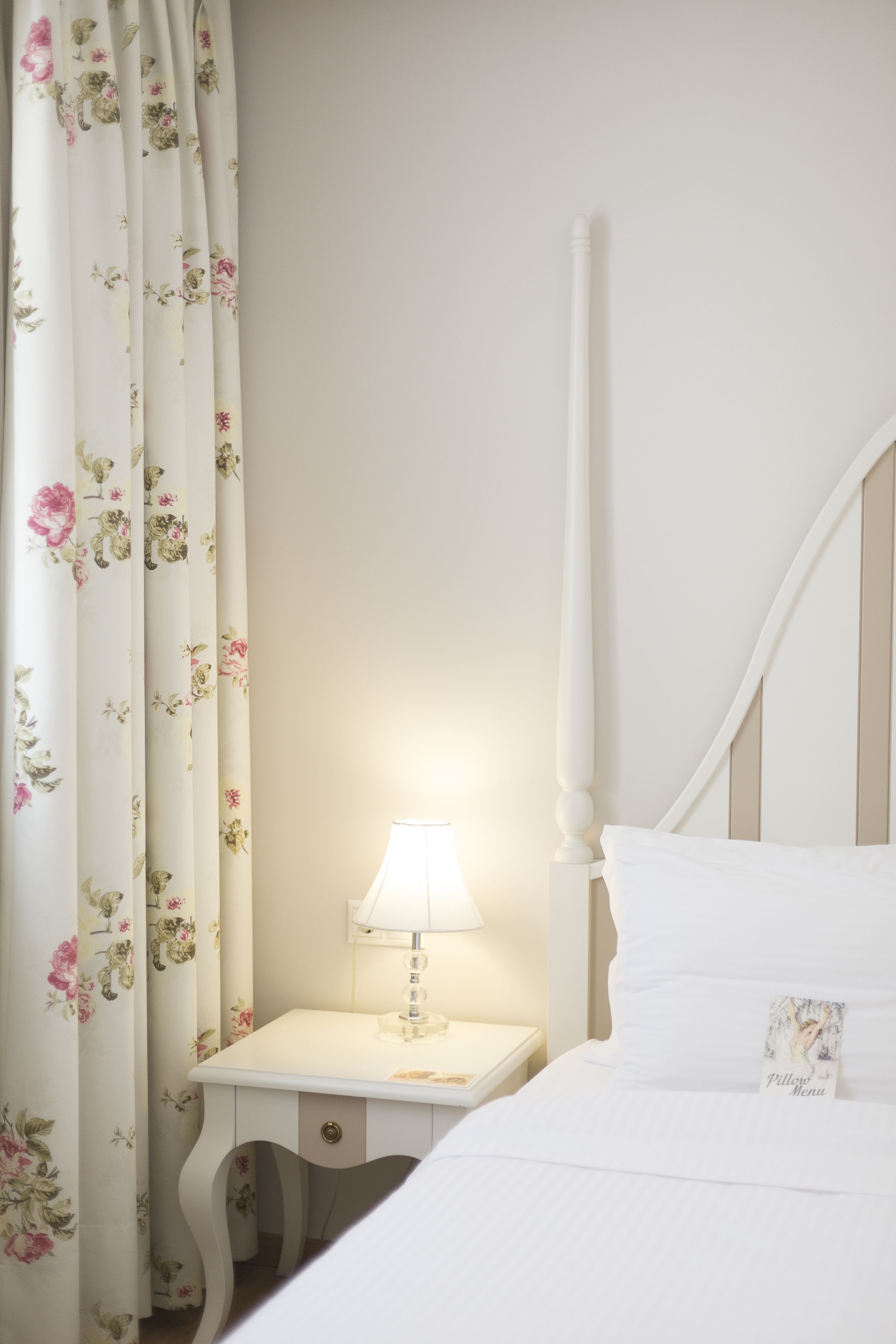 A neatly made bed with a pillow labeled "Pilote Menu," a bedside table with a lamp, and floral curtains.