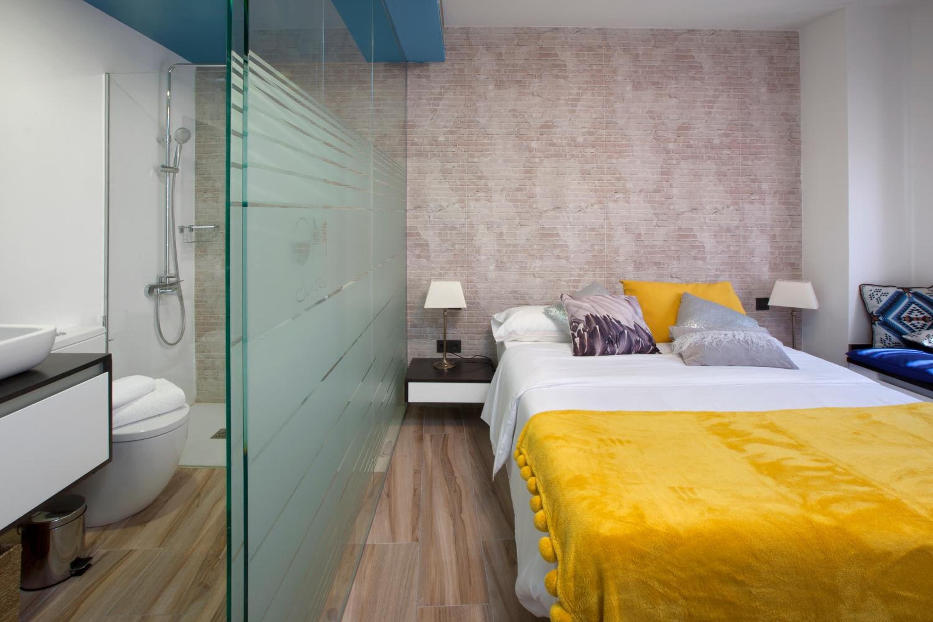 A modern hotel room with a glass shower enclosure, a bed with a yellow throw, and a brick-patterned wall.