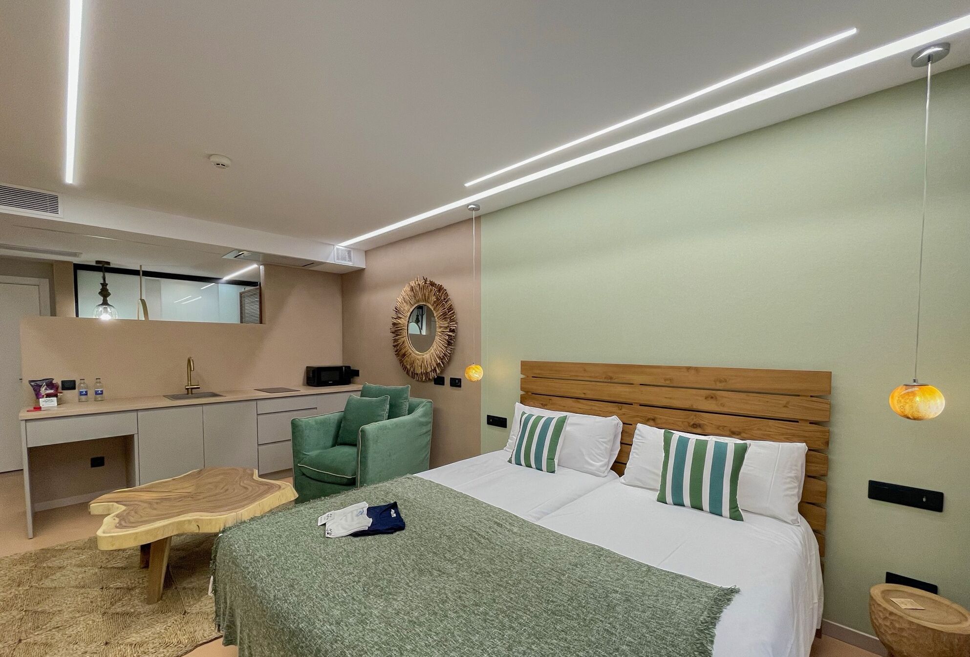Una camera d'albergo moderna con un letto, una testiera in legno, una poltrona verde, una scrivania con uno specchio e una lampada a sospensione.