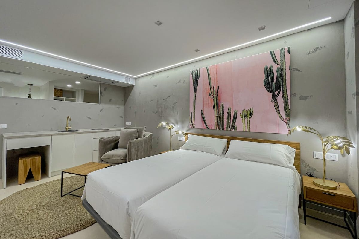 A modern bedroom with a double bed, a bedside table, a nightstand, a desk with a chair, a mirror, and a large cactus-themed wall art.