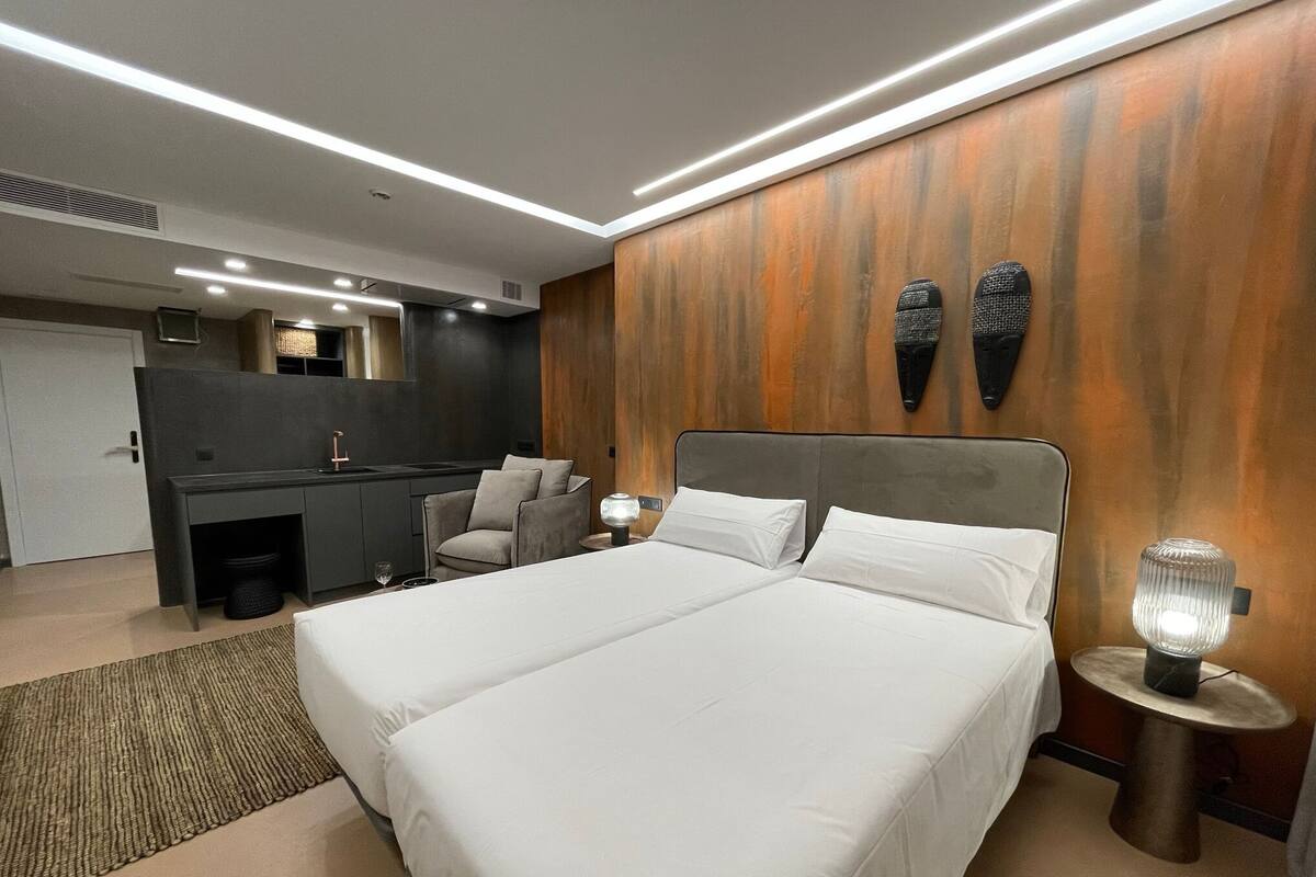 A modern hotel room with a large bed, a desk, a chair, and a small table with a lamp.