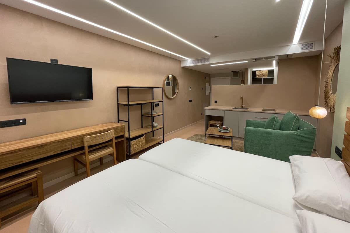 A modern hotel room with a large bed, a desk, a TV, a mirror, and a small kitchenette.